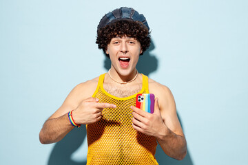 Young happy gay Latin man wear mesh tank top hat clothes hold use point on mobile cell phone isolated on plain pastel light blue cyan background studio portrait Pride day June month love LGBT concept