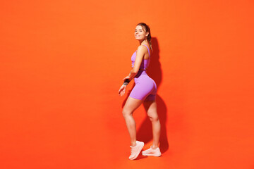 Full body side view young smiling fitness trainer instructor woman sportsman wear purple top clothes train in home gym look camera isolated on plain orange background. Workout sport fit abs concept.