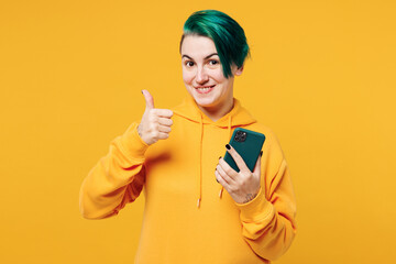 Young happy woman with dyed green hair wear hoody casual clothes hold in hand use mobile cell phone show thumb up gesture isolated on plain yellow orange background studio portrait Lifestyle concept