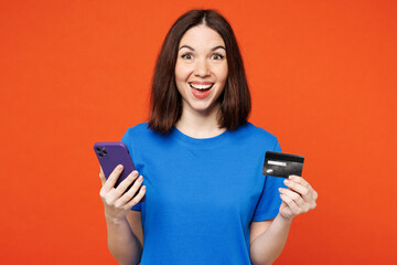 Young woman wear blue t-shirt casual clothes using mobile cell phone hold credit bank card doing online shopping order delivery booking tour isolated on plain red orange background. Lifestyle concept.