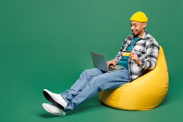 Full body young man wear shirt t-shirt yellow hat sit in bag chair using laptop pc computer hold...