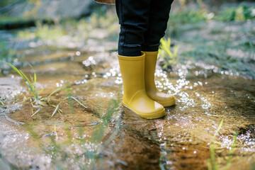 Feet of a small child in yellow rubber boots stand in a stream sparkling in the sun. Cropped....