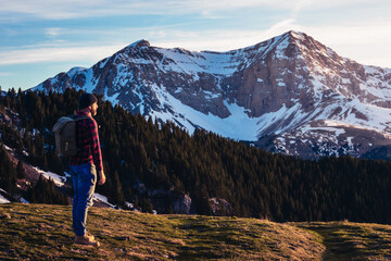 A man stands surrounded by high mountains and forests. The man carries a backpack and enjoys the view of the landscape.