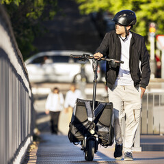 A delivery man from an urban delivery app with his skateboard and black delivery bag walking across...