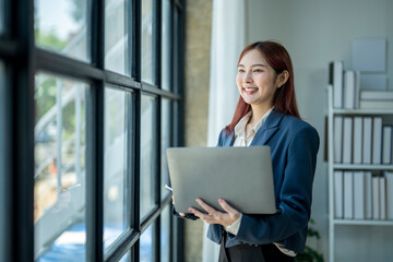 A woman in a business suit holding a laptop in front of a window. She is smiling and she is happy