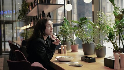 Young Caucasian beautiful woman with dark long hair and a pleasant smile sitting in restaurant. Girl is eating salad, and talking to her phone.