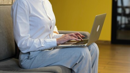  video. Businesswoman in white shirt and grey trousers is sitting on the grey couch and working on her computer, looking into the camera and smiling.. - Powered by Adobe