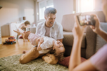 Father playing with daughter while being photographed by mother at home