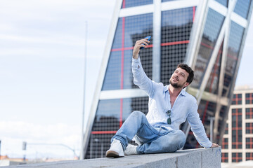 Man on vacation in the city taking a photo with his mobile phone. Happy and smiling man taking a photo with his cell phone. Boy sitting taking a photo with his cell phone of him in the city.