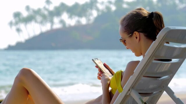 Brunette woman makes videocall to friend sitting on sunbed on ocean beach. Female donned in swimsuit sunbathes on ocean shore