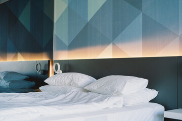 The scene captures a contemporary bedroom featuring a stylish geometric pattern on the wall. An unmade bed with rumpled white sheets and pillows sits in the foreground, suggesting a relaxed