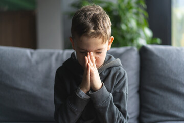 A young boy is seated on a soft sofa, his hands pressed together in front of him as if in is...