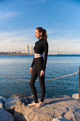 Young woman in a sports wetsuit posing on rocky seaside with city skyline in the background