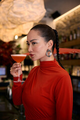 A sophisticated asian woman in a vibrant red turtleneck and detailed earrings holds a cocktail with a poised expression. She stands in a chic bar, with artistic lighting creating a warm ambiance.