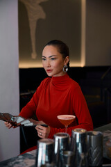 An elegant asian woman in a vibrant red turtleneck dress stands gracefully at a bar, holding a...