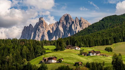 A valley of green meadows surrounded by pine trees, wooden houses and towering mountains. Vall di...