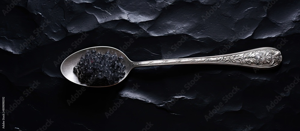 Poster Top view of a silver spoon holding black sturgeon caviar placed on a black slate stone background with enough space for adding text or other images. Creative banner. Copyspace image