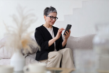 A mature woman relaxes on her sofa, using her smartphone with confidence and modern technology.