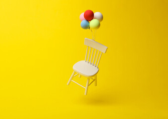 White miniature chair tied to balloons floating on a yellow background. Minimalism.