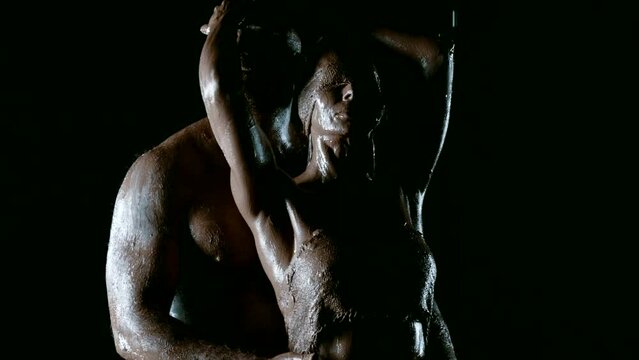 In the darkness on a black background, a stunning scene of passion and devotion unfolds, where a man with a muscular physique and his beautiful wife, decorated with clay