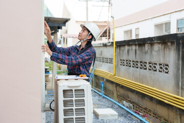 Asian woman in plaid shirt, white helmet, examines outdoor air conditioning unit with focus,...