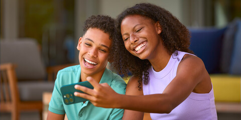 Teenage Brother And Sister Posing For Selfie On Mobile Phone Sitting Outdoors At Home On Deck