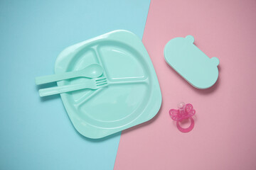 Set of plastic dishes for feeding baby and pacifier on a blue-pink background. Top view