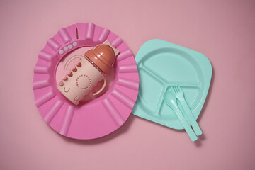 Set of plastic dishes for feeding baby, bib on a pink background. Top view. Flat lay