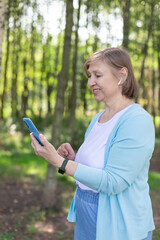 Beautiful smiling mature woman with wireless headphones, phone and smart watches walking in nature. Healthy lifestyle concept