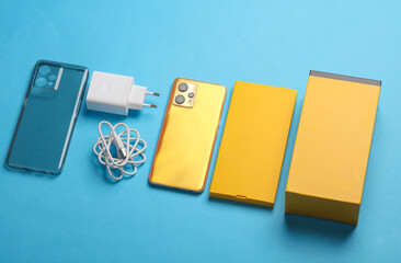 New yellow smartphone with box and accessories on blue background. Modern gadgets, unboxing. Flat lay. Top view