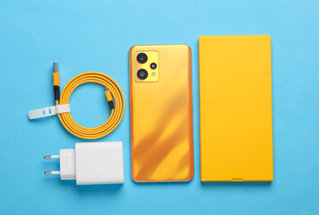 New yellow smartphone with box and accessories on blue background. Modern gadgets, unboxing. Flat...