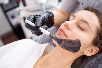 Female cosmetologist applying black mask on the face of a beautiful woman for carbon peel