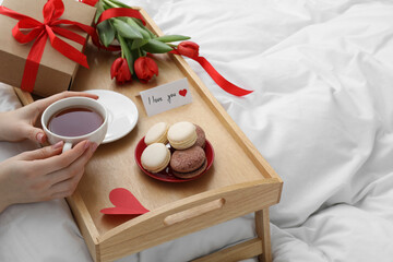 Tasty breakfast served in bed. Woman with tea, macarons, gift box, flowers and I Love You card at home, closeup