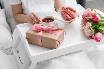 Tasty breakfast served in bed. Woman with tea, eclairs, gift box and flowers at home, closeup