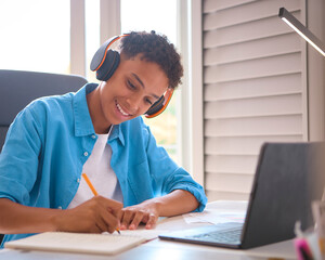 Teenage Boy Sitting At Desk In Bedroom At Home Doing Homework Streaming Music To Headphones