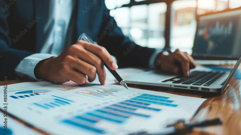Poster A person is examining financial graphs and data with a pen while working on a laptop in an office setting.