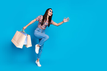 Full size photo of pretty young girl running hold shopping bags empty space wear trendy colorful clothes isolated on blue color background
