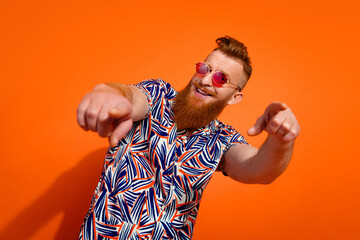 Portrait of optimistic positive man wear print shirt in glasses dancing indicating at you have fun isolated on orange color background