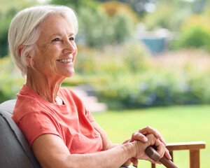 Independent Senior Woman Holding Walking Stick Sitting In Chair At Home Looking Out At Garden