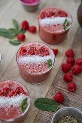A close-up view of vibrant raspberry smoothies, beautifully garnished with fresh mint leaves, coconut shavings, and juicy raspberry pieces. The refreshing beverages are served in clear glasses.