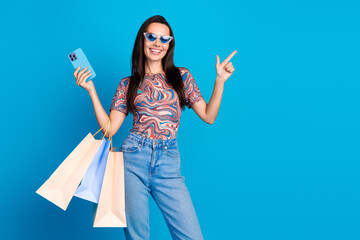 Photo of pretty young girl sunglass hold device shopper point empty space wear trendy colorful clothes isolated on blue color background