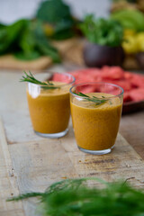 This image showcases two glasses of freshly blended green smoothie, garnished with rosemary, surrounded by an assortment of fresh vegetables and fruits like spinach and watermelon.