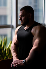 Pumped-up bodybuilder in a black T-shirt stands near the window in the gym. Handsome, strong, athletic rugged man pumping up his muscles. Training, sports nutrition, fitness and bodybuilding