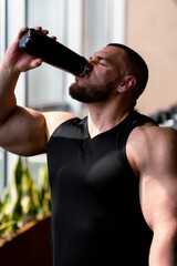 Pumped-up bodybuilder in a black T-shirt drinks water from a bottle near the gym window. Handsome, strong, athletic, strong man pumping up his muscles. Training, fitness and bodybuilding