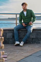 A young man in a green shirt and jeans sits casually by the sea, holding sunglasses and enjoying the serene waterfront view.