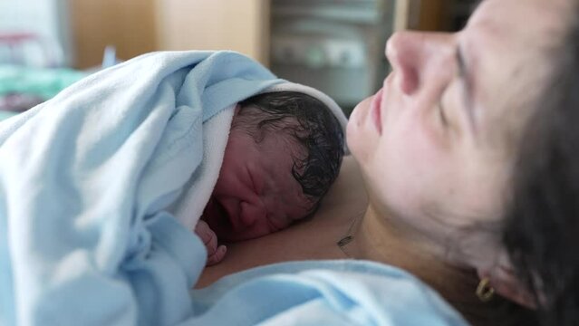 Crying newborn baby resting on mother's chest at hospital clinic right after birth, real life authentic childbirth scene of new infant arriving to this world