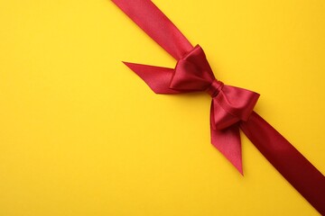 Red satin ribbon with bow on yellow background, top view. Space for text