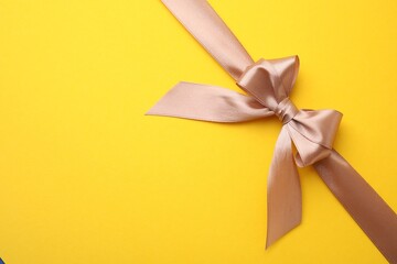 Beige satin ribbon with bow on yellow background, top view. Space for text