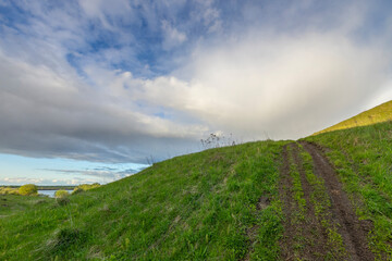 A tranquil dirt path on a grassy hill leads the eye towards a serene lake and a horizon filled with clouds, offering a peaceful and solitary outdoor experience.