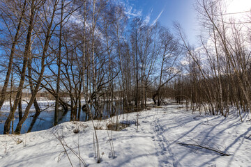 A serene winter scene with snow-covered ground and bare trees, sunlight casting shadows, a river reflecting the blue sky, and footprints leading along a path